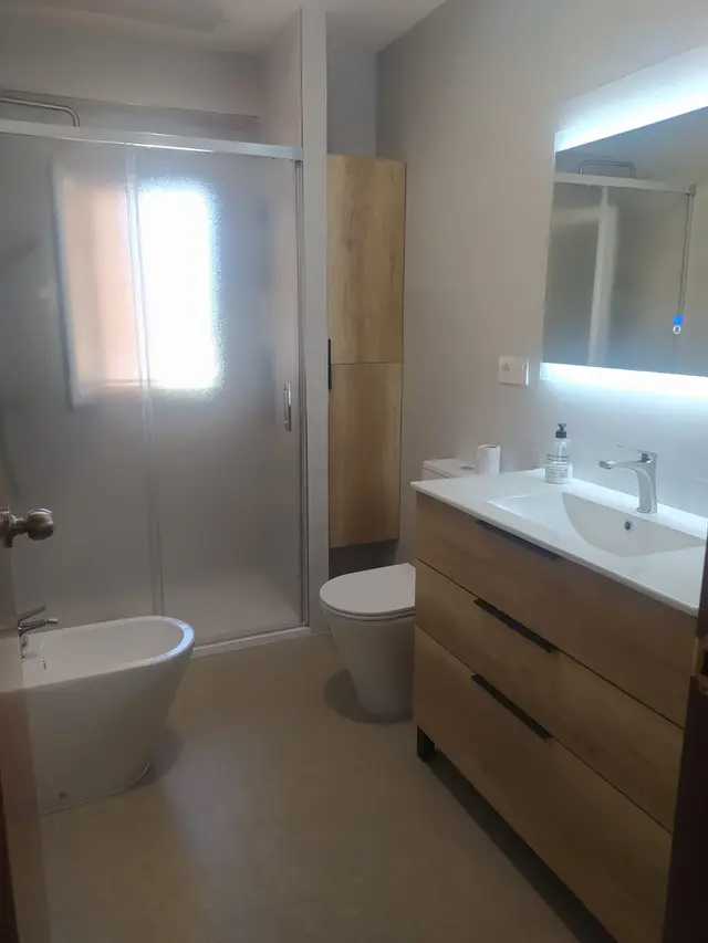 Baño con mueble de madera y espejo iluminado, trabajo de Dos Ambients